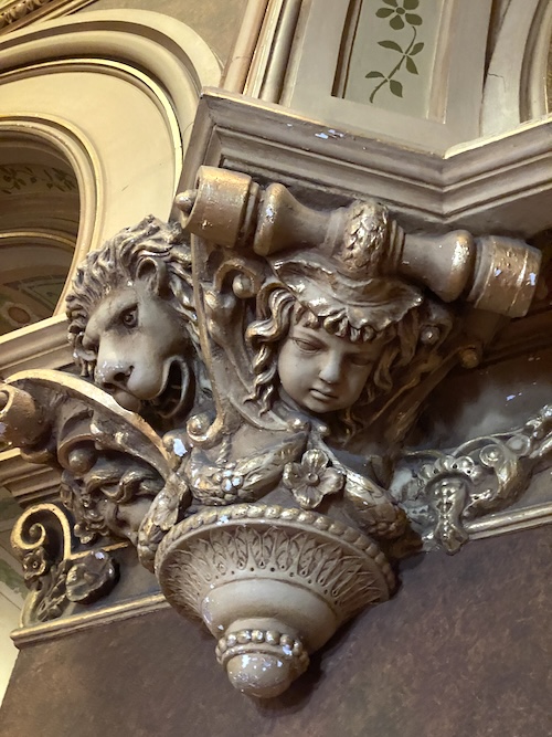 Photo of decorative top of column with a lion's face on the left and a girl's face on the right next to it.