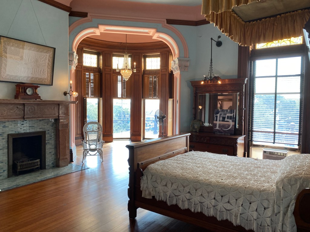 Photo of bedroom. Bed is covered in a lace bedspread, floor is light hardwood, fireplace mantle and sides are light wood and carved, fireplace has light grey/green tiles. Antique clock on fireplace. Beautiful wooden dresser with mirror sits next to the window. Small white wicker rocking chair sits in front of arched entry to a circular sitting area with floor to ceiling windows, wooden shades, and small chandelier.