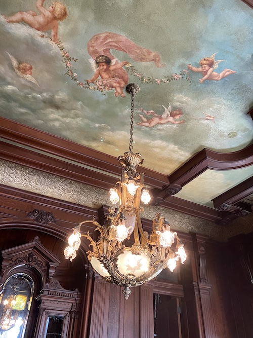 Photo shows ceiling painted with blue sky and clouds and cherubs, painted by Josephine Gresham. Surrounded by mahogany ceiling beams. Lace-looking wallpaper below the beams for about one foot, then mahogany walls with intricate carvings. Beautiful chandelier made of gold or brass with lights that are also intricate and look like flowers.