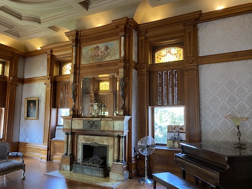 Photo shows room with a piano, fireplace with stone surrounding it, window above with carved pieces leading to more wood decor up to the tall ceiling. At the top is a painting of clouds, blue sky, and cherubs (painted by Josephine Gresham). On both sides of the fireplace are windows with upper wooden shades, and on top of them are short stained glass windows of yellow and white with a a bow design on top with ribbon leading to the sides of the window with another bow and the ribbons coming down. Gray wallpaper beside the windows, with diamond designs with leaves in the center of each diamond. 