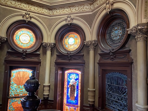 Photo is another view from the second floor showing the two stained glass windows (one iwth orange, yellow, and teal colors, the other with golden decor around a blue backgroud with a woman in the center) and also shows to the right of that window a darker window frame with gray designs in it (unsure if it is a light window as it looks dark from this view). Above these 3 windows are three circle stained glass. The first one on the left has colors matching the first stained glass window, the middle one has the same colors but styled a little differently, the third one is darker with some blue in it. 