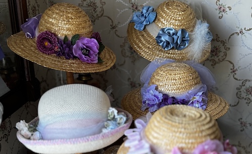 Hats on display. One white hat with pink and purple accents, four straw hats, one with pink flowers, one with blue bows and feathers, one with purple flowers and material, one with pink flowers and material.
