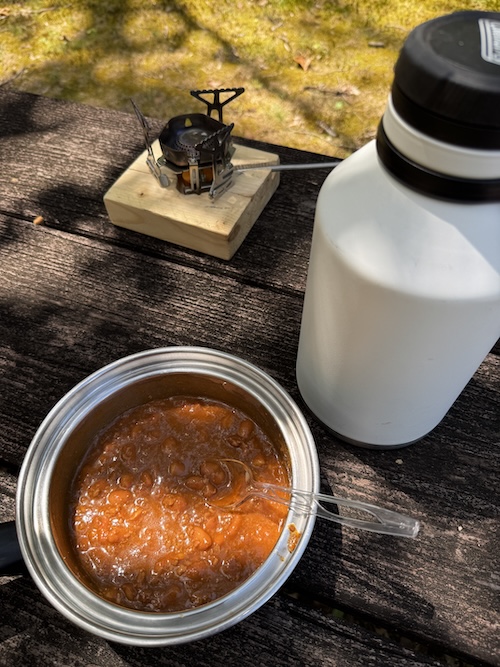 Image of a small cookstove in the upper part, a white thermos on the right, and a cookpot with chili and a spoon in it towards the bottom of the photoe.