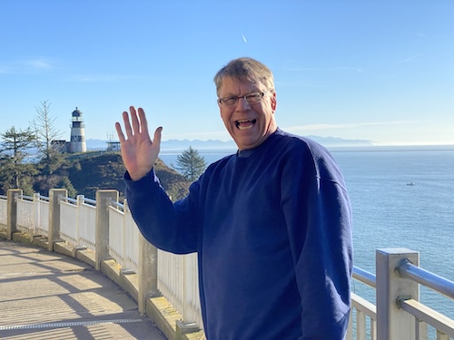 David waving, standing outside in front of high view of ocean, with a lighthouse in the background.