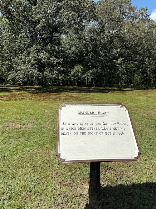 Small sign saying, "Grinder House. Site and ruins of the Grinder House, in which Meriwether Lewis met his death on the night of October. 11, 1809. In the background is a grassy field with trees at the back.