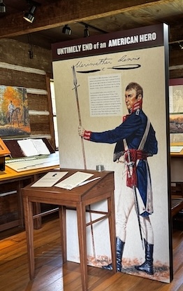 Image of Meriwether Lewis inside of visitor center. He is wearing a blue military jacket, white pants, black boots, and holding a staff of some sort. In front of picgture is a small wooden desk with documents on it. In background is more documents and a picture on the wall of a man on horseback riding through trees with orange leaves.