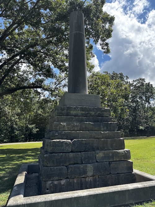 Image of Meriwether Lewis monument made of base, oblong blocks on top of each other, narrowing as they go up a couple of feet, then a cylindrical column. Grassy field with trees in the background.