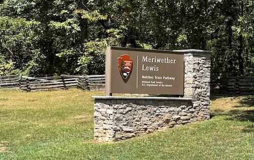 Image of National Park sign saying Meriwether Lewis, Natchez Trace Parkway" on brown board with National Park emblem on top of rock base. Grassy area with old fence and trees in background.
