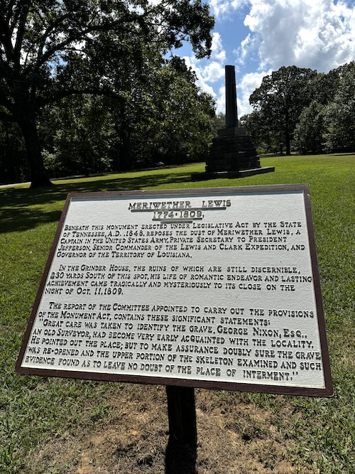 Image of Meriwether Lewis informational sign in front of tall monument to Lewis. Green grassy field, trees in the background.