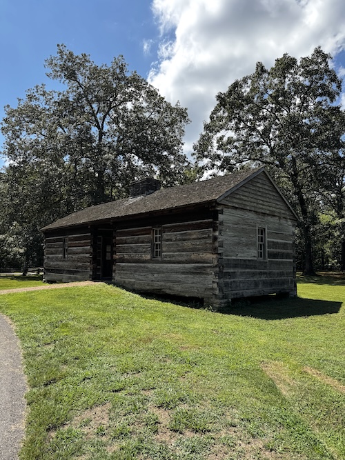Image of small old-looking log building sitting on a grass lot with trees in the background.