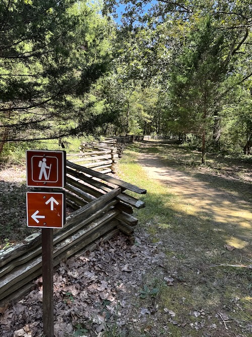 Image of trail,surrounded with trees on both sides, wooden fence on the left side, and hiker sign in front.